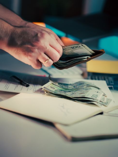 Close-up of a person's hand holding a worn wooden wallet with a transparent window, positioned over a surface cluttered with various banknotes including British pounds and euros, some bills partially 