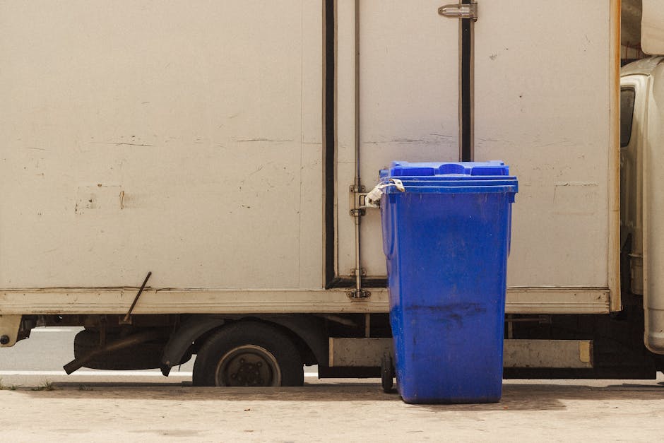 A blue waste bin positioned outside a white, metal food delivery van, which is parked on a concrete surface. The truck's side panel appears slightly worn with some dirt and scratches, and the bin is s