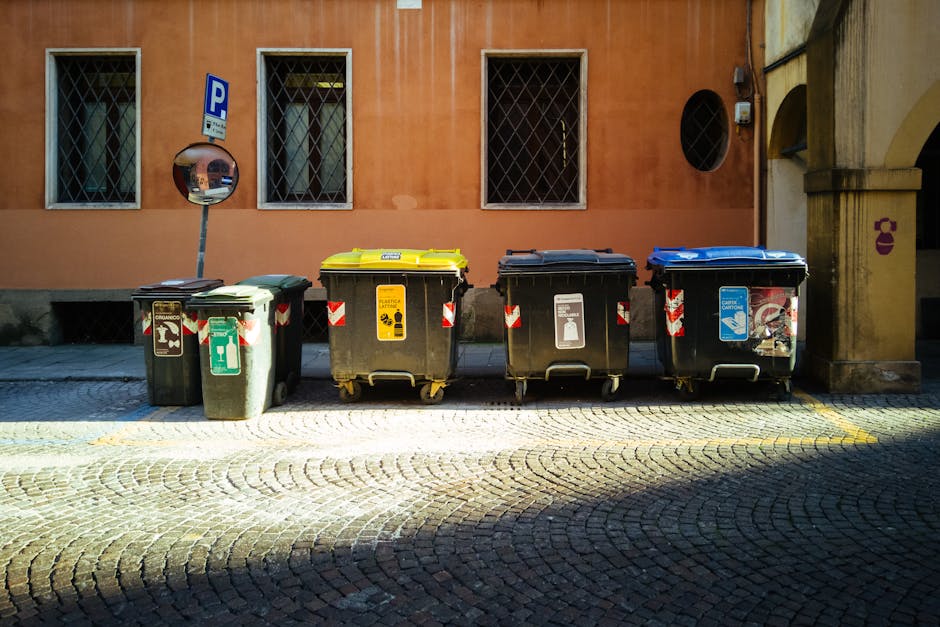 A blue waste bin positioned outside a white, metal food delivery van, which is parked on a concrete surface. The truck's side panel appears slightly worn with some dirt and scratches, and the bin is s